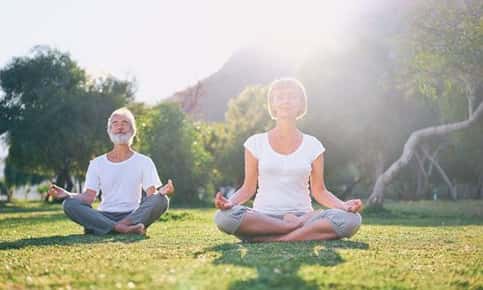 Image of two seniors seated in a yoga position outside in the park. 