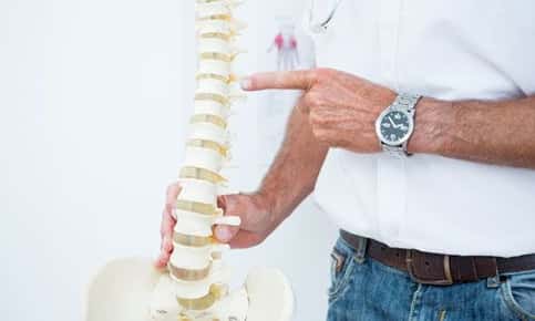 Image of a male chiropractor in his clinic pointing to a replica human spine to educate a patient. 