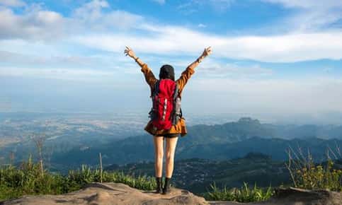 An image of a female backpacker who is standing on top of a mountain, in a victorious stance with both of her arms raised high.