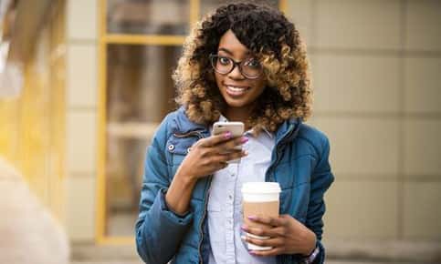 Image of women wearing glasses who is outdoors with her cellphone in one hand while holding coffee in the other. 