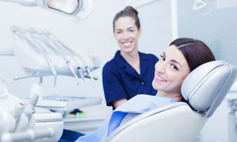 An image of a smiling female dental patient lying in the exam chair. A female dental hygienist is sitting beside the patient, smiling as well. 