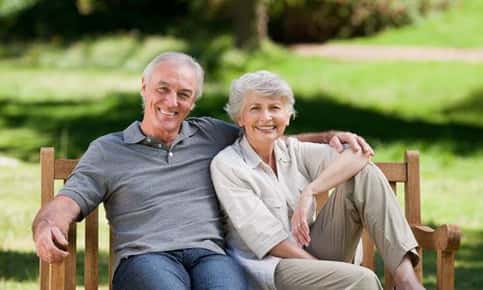 An image of a retired couple sitting on a park bench and smiling. 