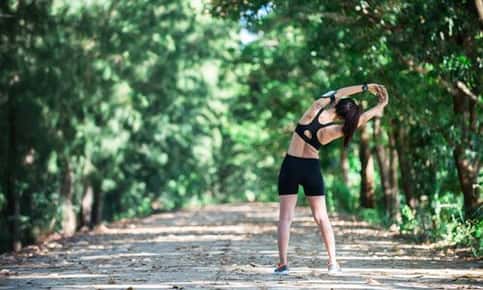 An image of a woman doing a stretch with her hands clasped and her arms stretched upwards. She's wearing workout attire while on a shaded trail surrounded by trees. 