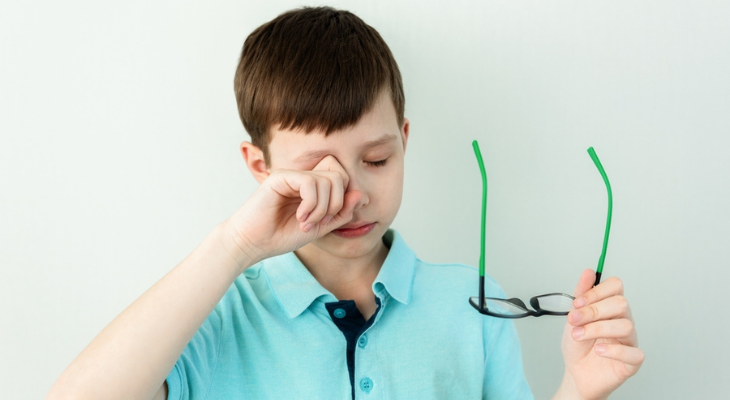 Boy rubs his eye while holding glasses.