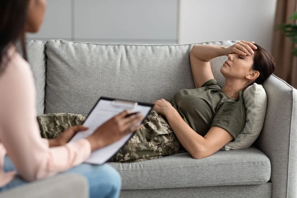 Woman wearing camouflage fatigues laying on a couch with her hand on her head, talking to therapist