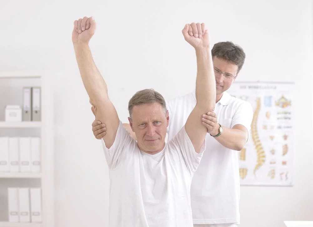 Chiropractor helping patient relieve his joint pain by properly stretching his arms over the top of his head