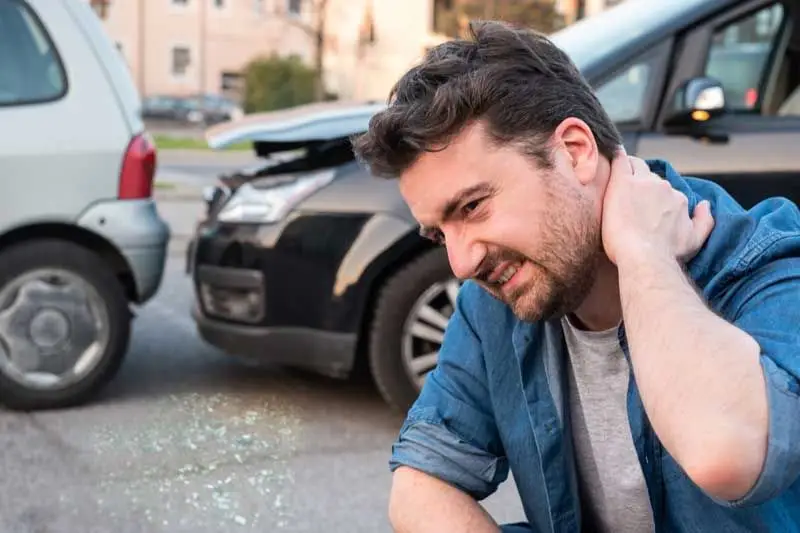 man kneeling down between 2 wrecked cars holding his neck