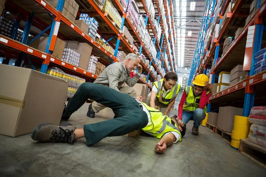 A worker on the floor in a warehouse. There are boxes on the floor and three other employees attending to the worker.