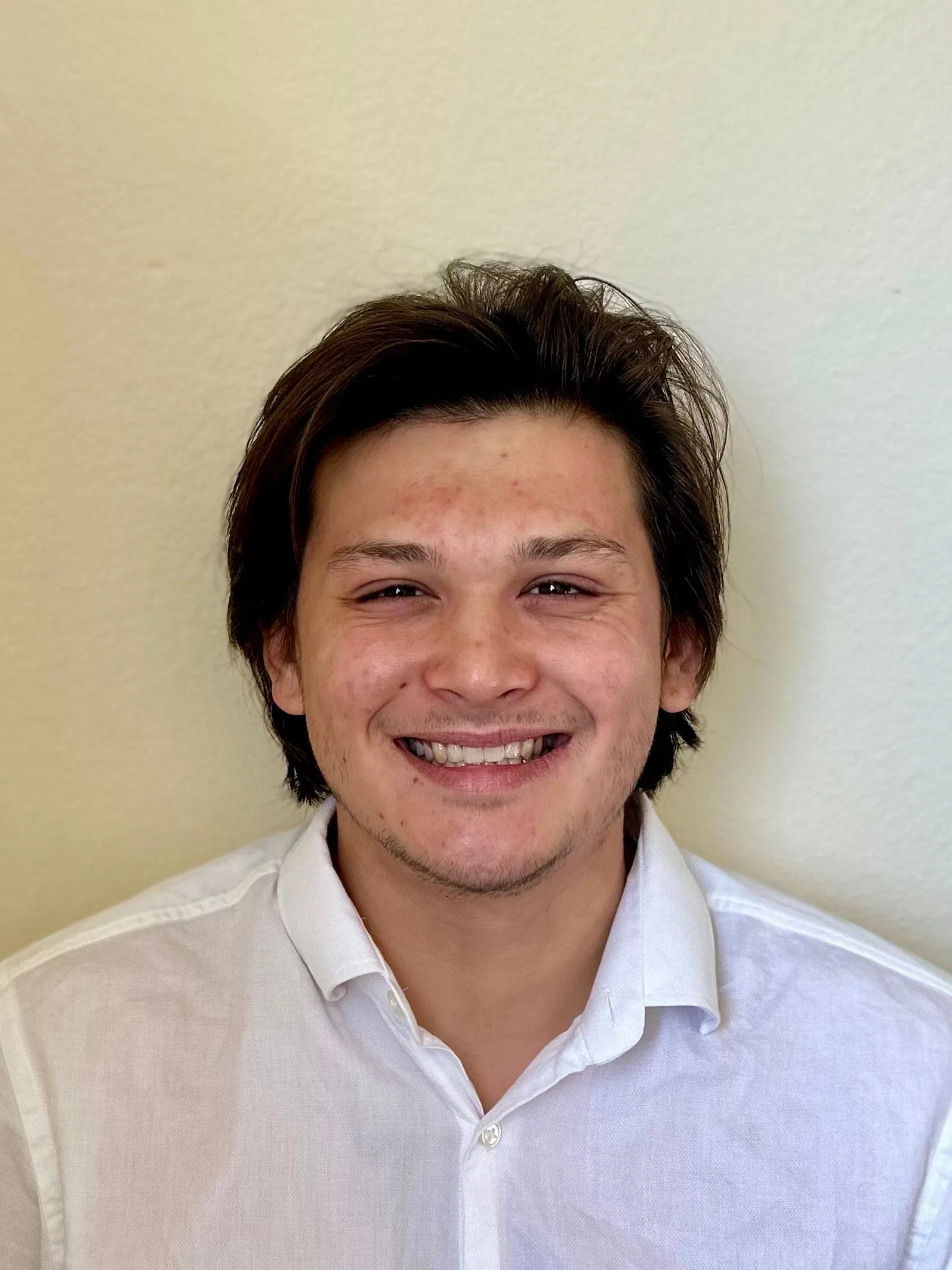 Gabriel stands in front of a blank wall in a white shirt, smiling.