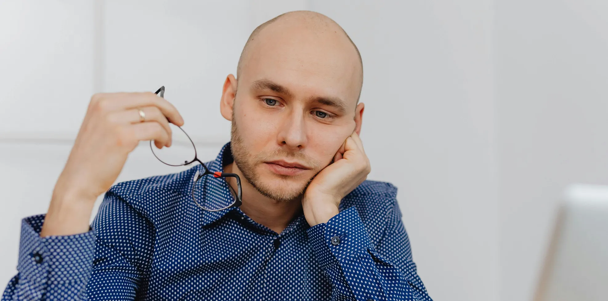 man sitting by a computer holding his glasses