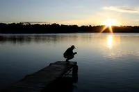 man squatting on edge of pier on a lake at sunset