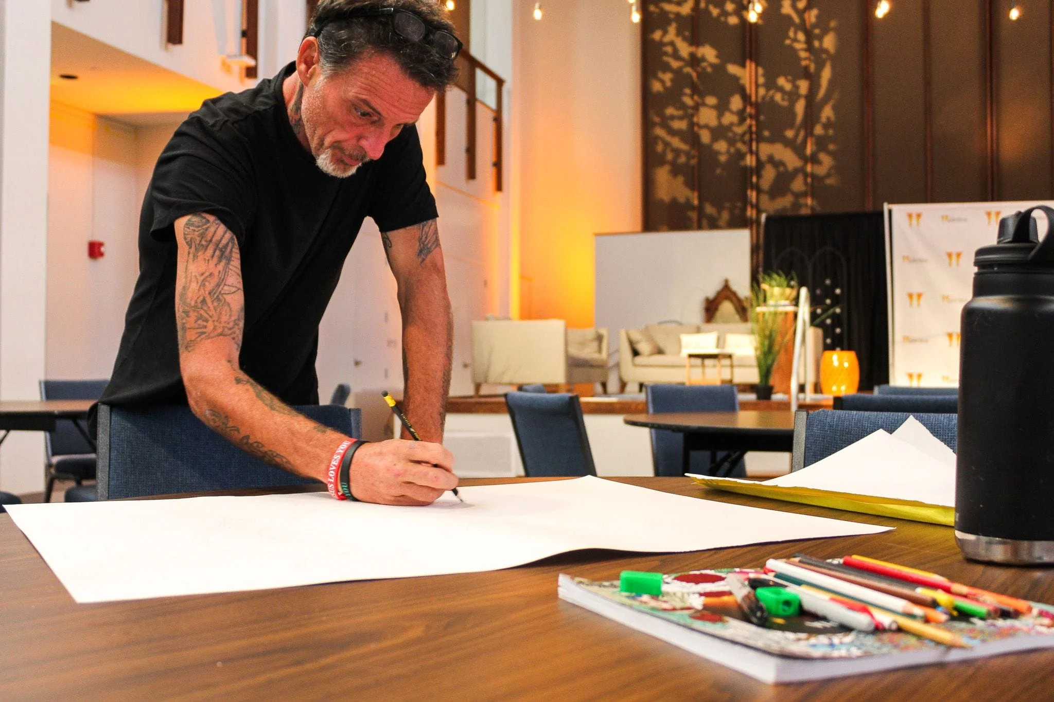 A man drawing on a large paper with art supplies on the table, and ambient lighting in the background.