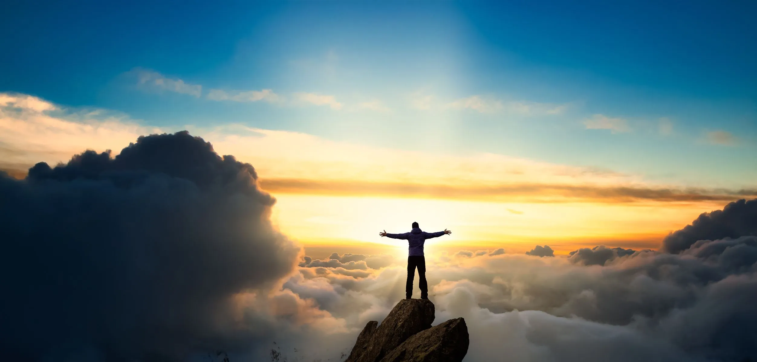 Man raising hands on top of a mountain above the clouds facing the sunrise.