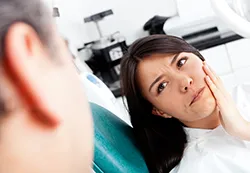 Asian woman sitting in dental exam room holding side of jaw in pain, toothache, needs root canal South Orange, NJ family dentist