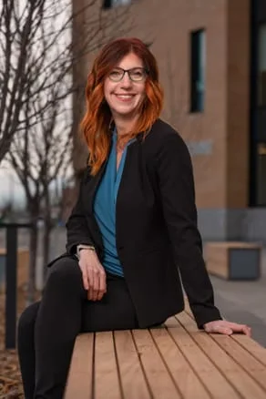 Owner of Wright Step Coaching, Caucasian female with red hair wearing a black suit, blue shirt sitting on a wooden bench outside on a fall day.