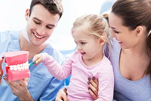 male dental hygienist showing toddler girl model of teeth while mom holds her, pediatric dentist Baytown TX