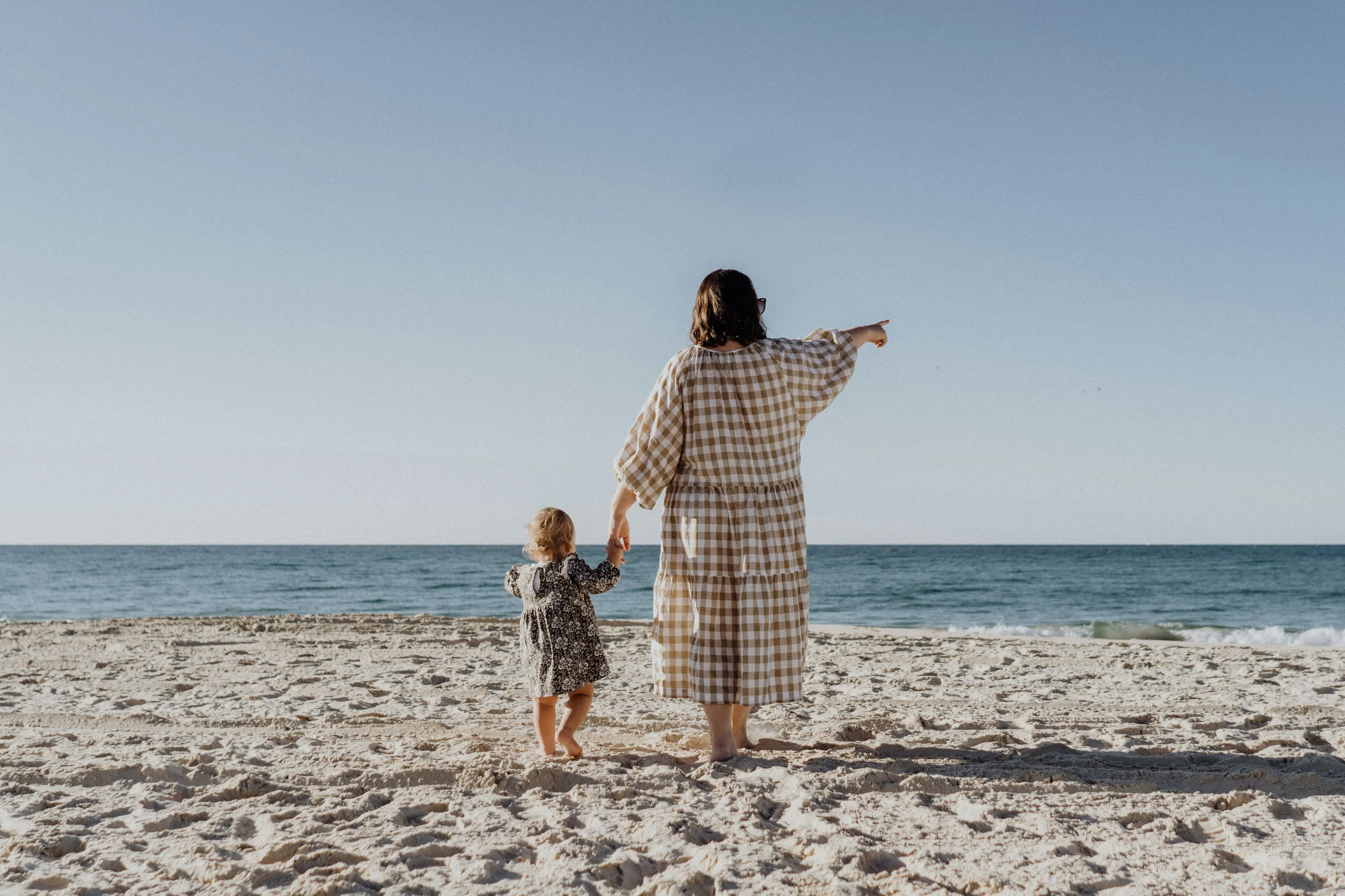 Woman holding child's hand on the beach pointing to the ocean