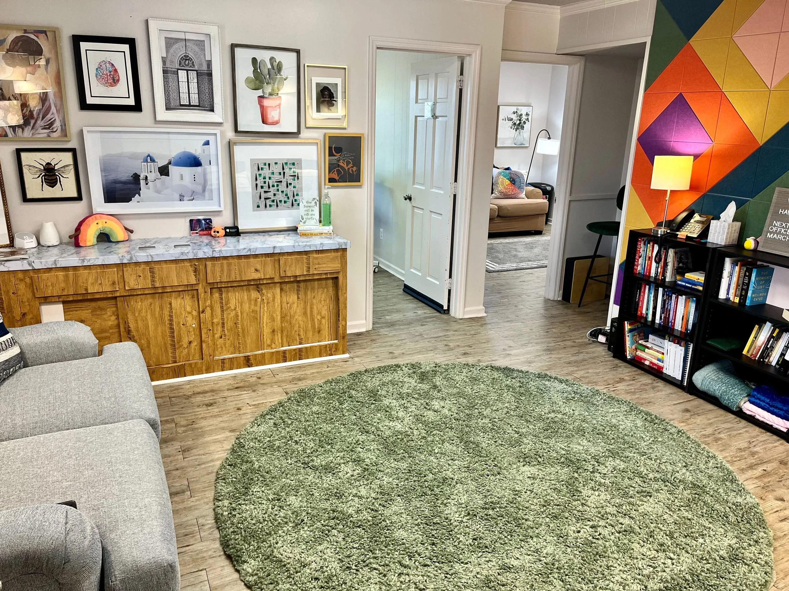 picture of office from entry door; waiting room with collage photo wall, gray couch to left, green circle rug, black bookshelves on right wall. There are two offices