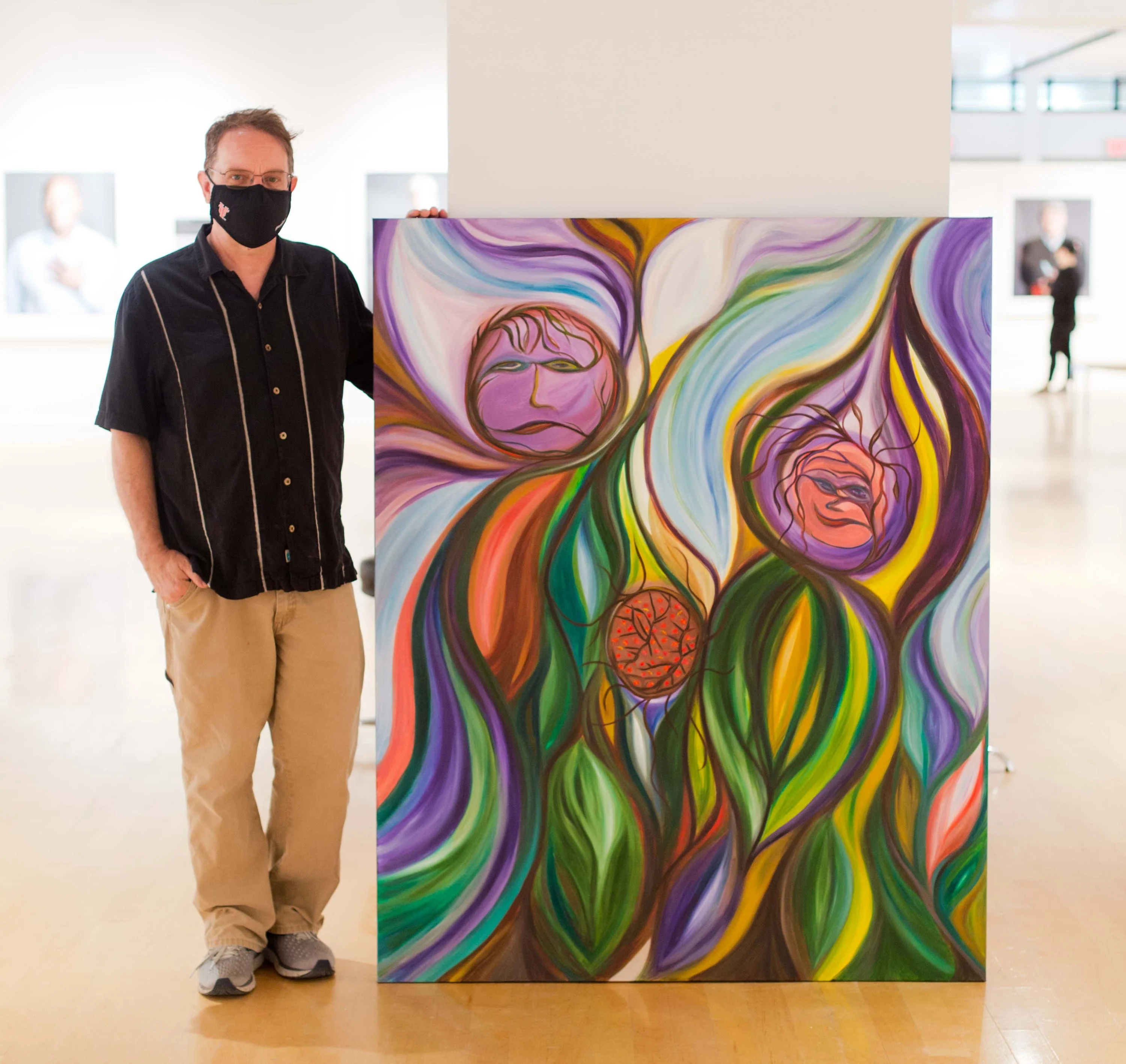 A man standing next to a large painting, the painting goes up to his chin and depicts blended colors and organic shapes, with three abstract faces.