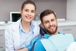 female dental hygienist smiling posing next to male patient in dental chair wearing dental bib, family dentistry Newark, CA