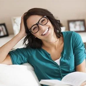 A woman wearing glasses holding an open book while sitting on a sofa and smiling