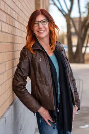 Photo of the owner of Wright Step Coaching. Tall thin woman with red hair, wearing a brown leather jacket, jeans and a blue shirt 