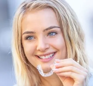 head of blond teen girl smiling, holding clear aligner tray in left hand, Invisalign Montclair, NJ dentist