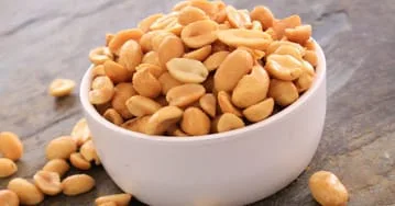 A bowl of peanuts on a wooden table. Some nuts are spilled across the table