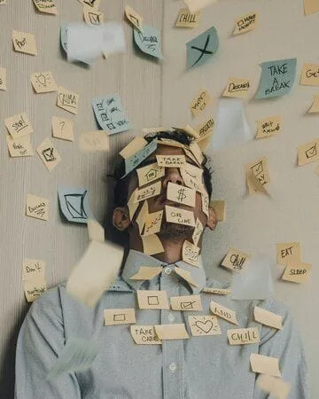 A person with short brown hair in a pale blue button-down shirt leans back into a corner, surrounded and covered with sticky notes.