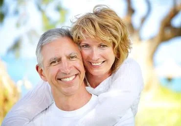 older man and woman hugging, smiling nice teeth, outdoors near trees and ocean, dental implants San Diego, CA dentist
