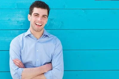 college age man in button up shirt leaning against blue wood wall, smiling nice white teeth, family dentist Gardnerville, NV