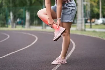 Female athlete standing on track, clutching ankle and lower leg in pain from Achilles injury