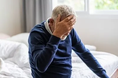 Senior man holding his head in pain sitting on bed.