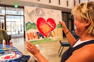 Image of a woman holding up her painting and smiling at it. The painting depicts a heart and grass, and reads, "hope, healing, humanity."