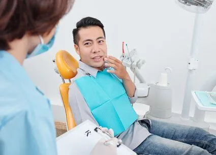 man sitting in dental exam chair gesturing to mouth in pain, talking with dental assistant, emergency dental care Washington DC dentist