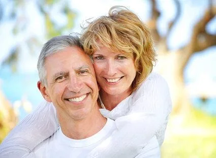 older couple hugging, smiling nice teeth, outdoors by trees and lake, dental implant dentist Beverly Hills, MI