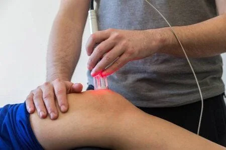 a person performs laser therapy on a patient's knee with a white wand emitting a red light.