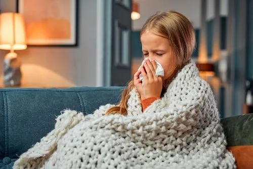 Child with flu covered in blanket, sneezing on a sofa.