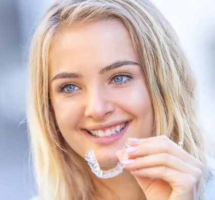 head of blond teen girl smiling, holding clear aligner tray in left hand, Invisalign Montclair, NJ dentist
