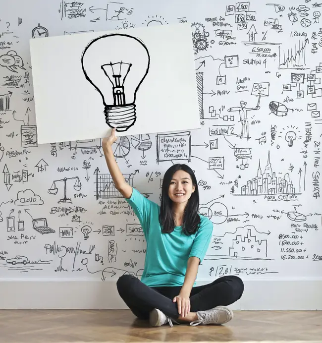 Girl sitting on the floor holding a picture of a light bulb. On the wall behind her is an abstract math equation