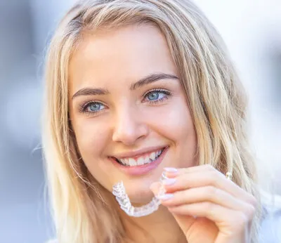 blond teen girl smiling, left hand holding clear aligner tray near mouth, Invisalign St Petersburg, FL dentist 