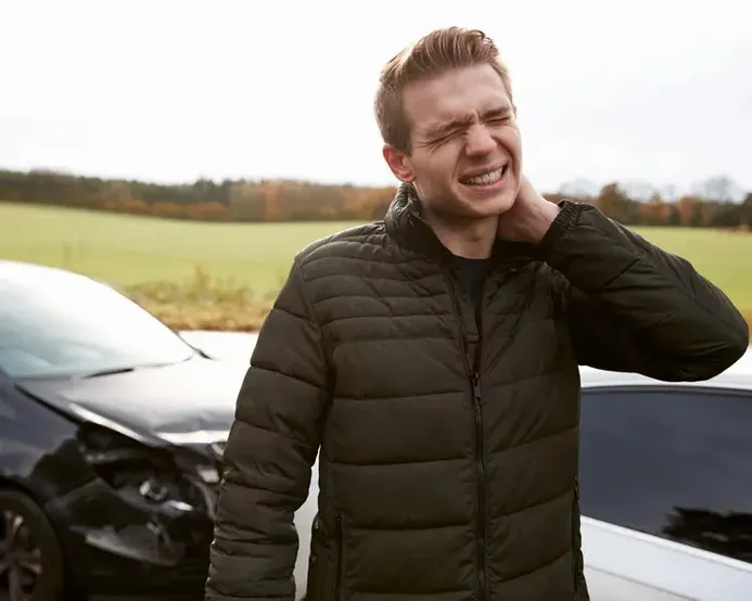 A young man with his hand on his neck wincing in pain. A car accident is behind him.