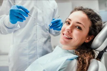 young woman smiling wearing dental bib in exam chair, dentist in background holding dental tools, dental cleaning Truro, NS