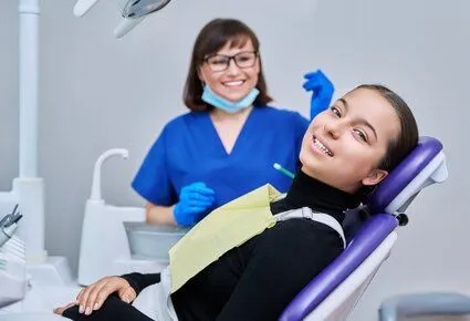 girl lying back in dental chair smiling, hygienist in background preparing to clean teeth, general dentist Livonia, MI 