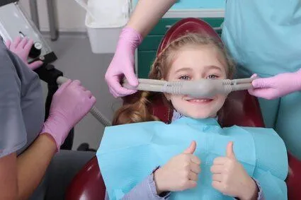 young girl sitting in dental chair giving thumbs up, nitrous oxide mask over nose, sedation dentistry North York, ON