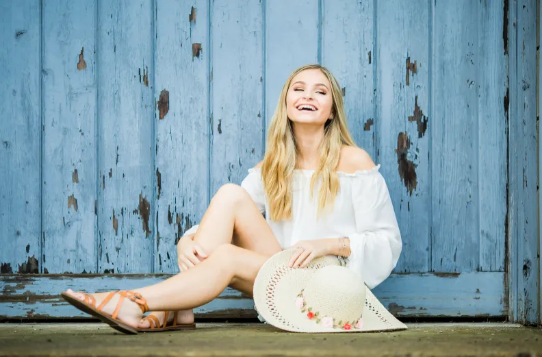 blond teen girl in white dress and sandals laughing, sitting in front of blue wood wall, family dentist Gardnerville, NV