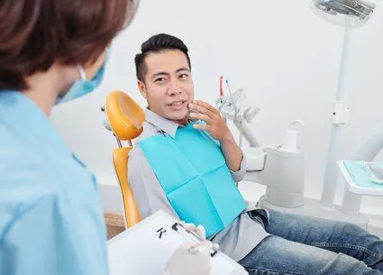 Asian man sitting in dental exam chair holding hand to mouth in pain, talking with dentist, root canal Sachse, TX dentist