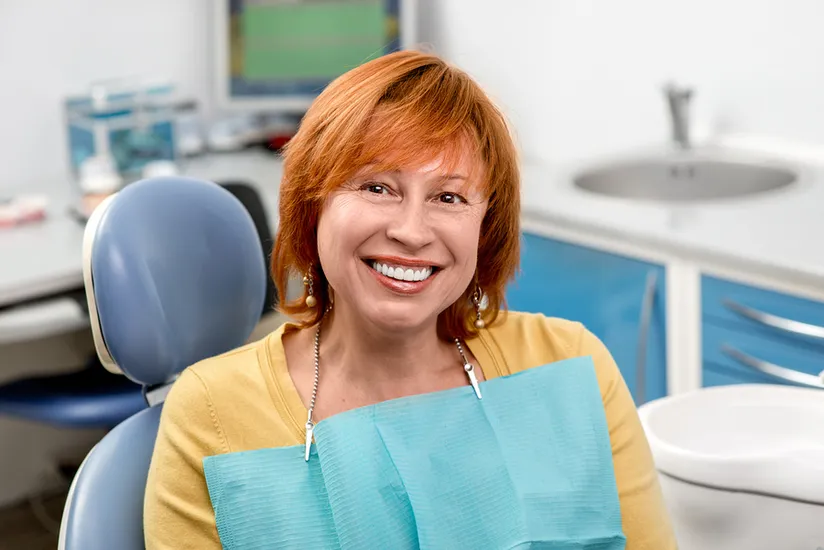 older red haired woman smiling, sitting in dental chair wearing dental bib, dental implants Peachtree City, GA