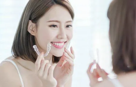 young woman holding clear aligner tray looking in mirror at teeth, Invisalign Brookline, MA dentist