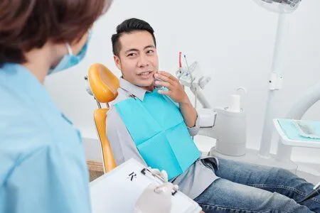 man gesturing to mouth in pain, sitting in dental exam chair looking at female assistant, root canal Boca Raton, FL dentist